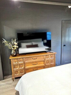 A bedroom with a bed, a wooden dresser with drawers, a flat-screen TV on top, and a vase with flowers on the dresser. - Davis St Apartment Complex | Whole House Near SFA (Nacogdoches)
