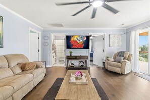 A living room with a beige sofa, a coffee table with a vase and flowers, a TV displaying 'Tom and Jerry', and a ceiling fan. - White River Retreat Escape - Hot Tub, 5 BRs, 2 Homes (West Fork)