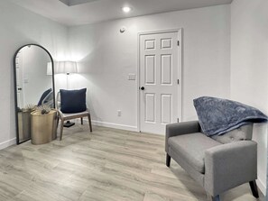 Una habitación con piso claro, una puerta blanca, una silla con cojín azul y una lámpara.