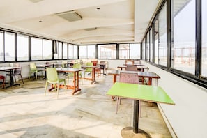 A spacious indoor area with multiple tables and chairs, large windows, and a high ceiling. - Collection O Infocity Metro Station (Gandhinagar)