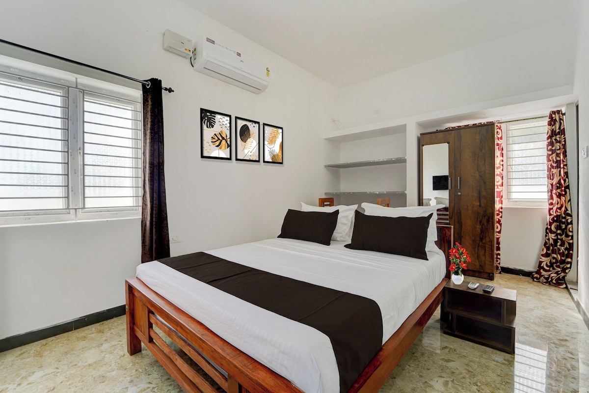 A hotel room with a bed, a window with blinds, a wall-mounted air conditioner, and three framed pictures.