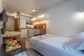 Ein kompaktes Hotelzimmer mit Bett, Schreibtisch mit Computer, einer kleinen Küchenzeile und einem Fernseher.
