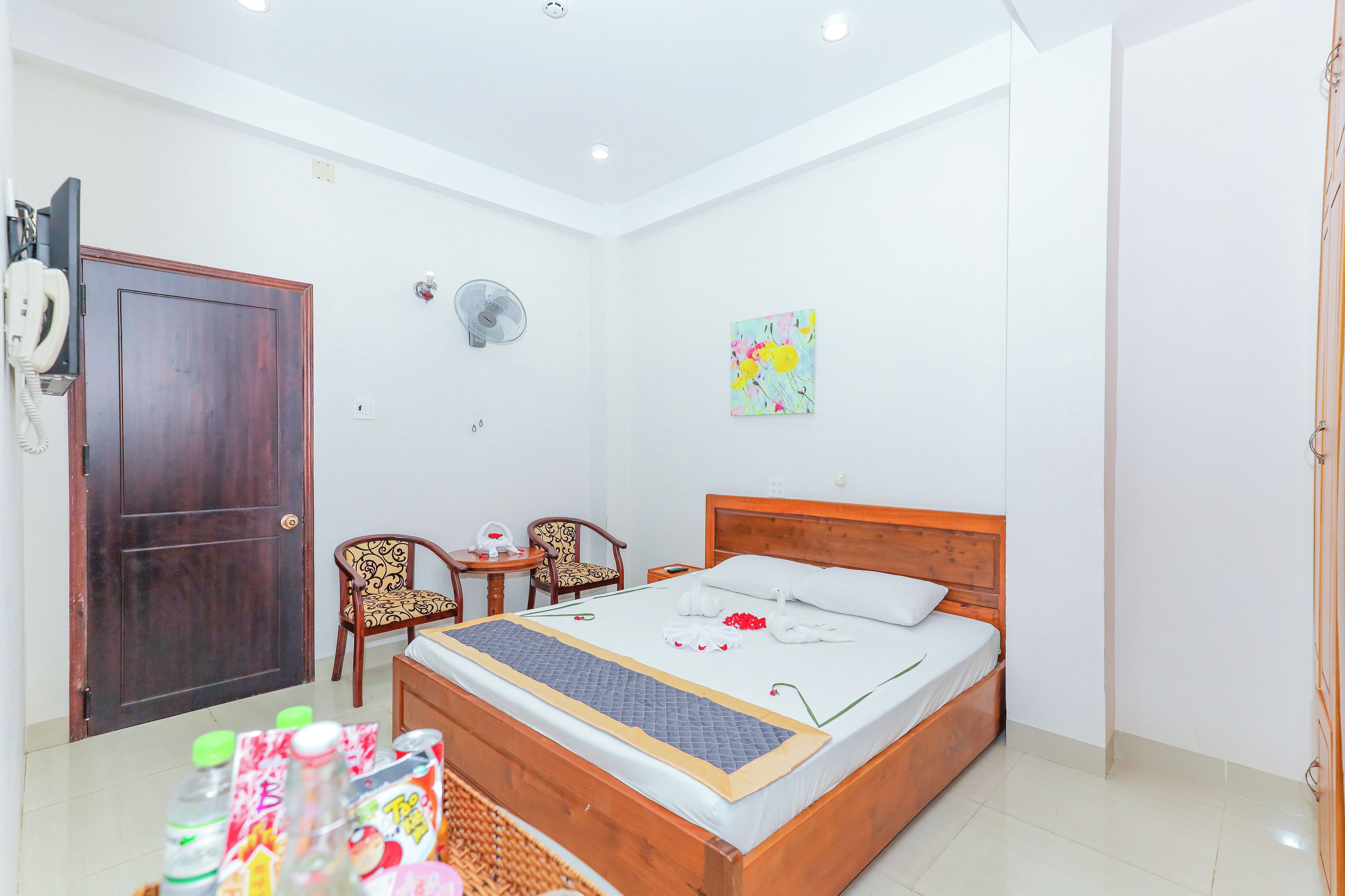 A hotel room with a wooden bed, a desk with a chair, a wall-mounted fan, and a door with a telephone.