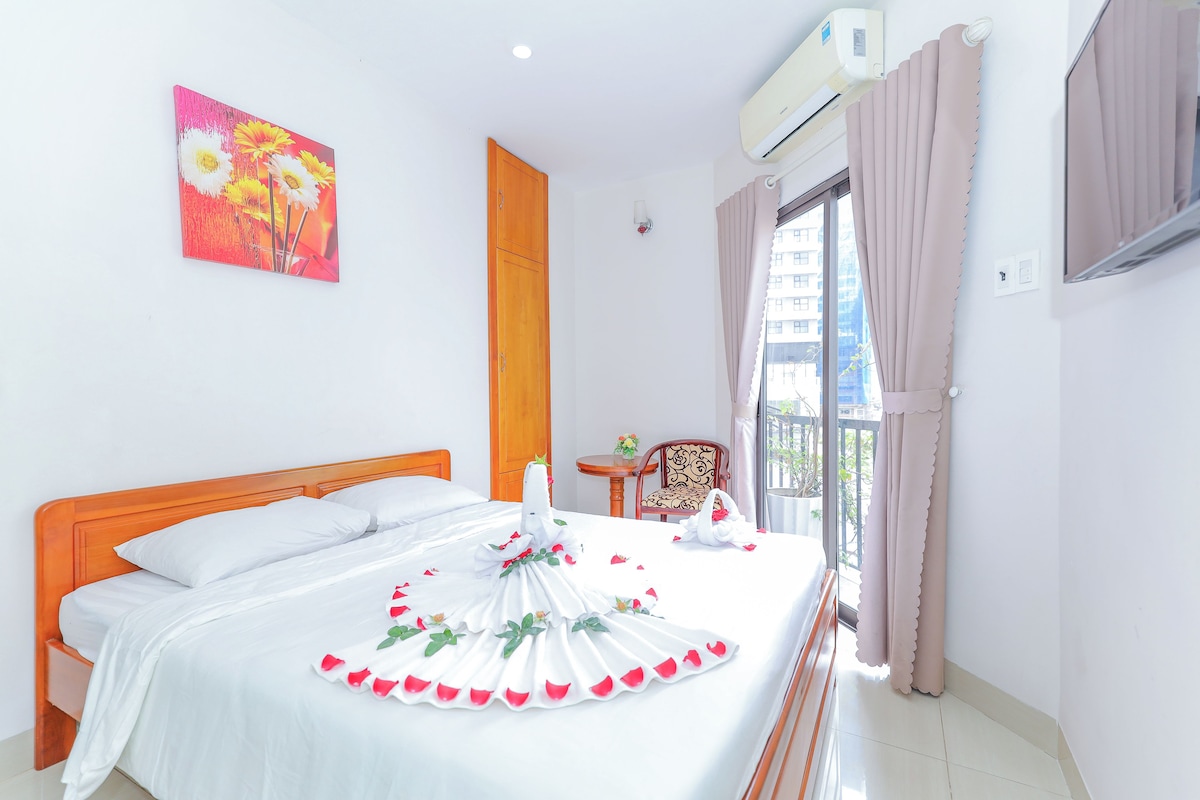Een hotelkamer met een bed, een houten kledingkast, een tafeltje met een vaas en een raam met gordijnen.