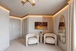 Quarto de hotel com um espelho grande, duas poltronas, uma mesa pequena com um vaso e um quadro na parede.