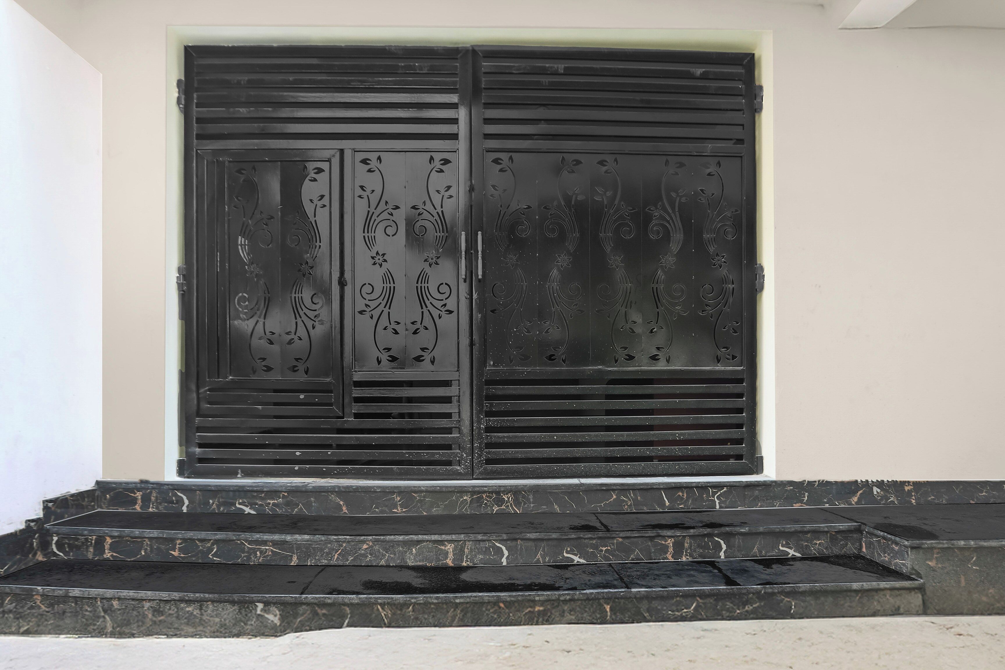 Una reja de metal negro con diseños decorativos, ubicada frente a una pared blanca, con escalones que conducen hacia ella.