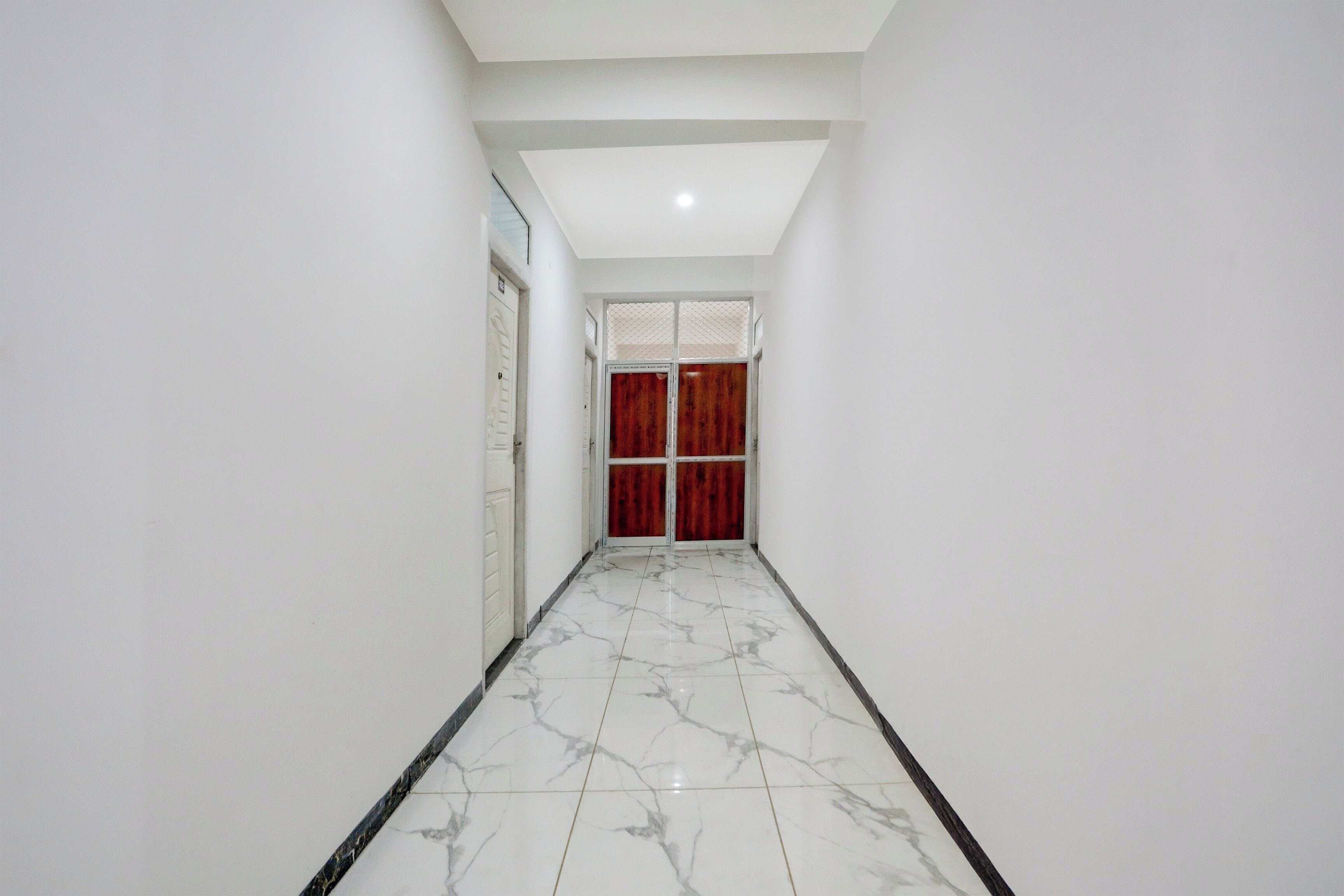 Um corredor com paredes brancas, chão de mármore e uma porta vermelha no final.