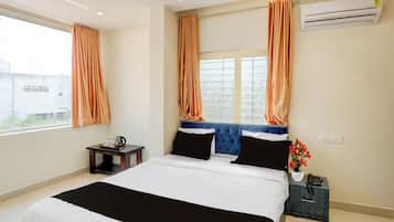 Een hotelkamer met een bed, een klein tafeltje met een telefoon en een bloemenvaas, een airconditioner en gordijnen.
