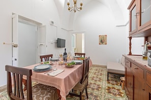A dining area with a table set for four, chairs, a chandelier, a television mounted on the wall, and a patterned floor. - Casa Giselda by BarbarHouse (San Giorgio Ionico)