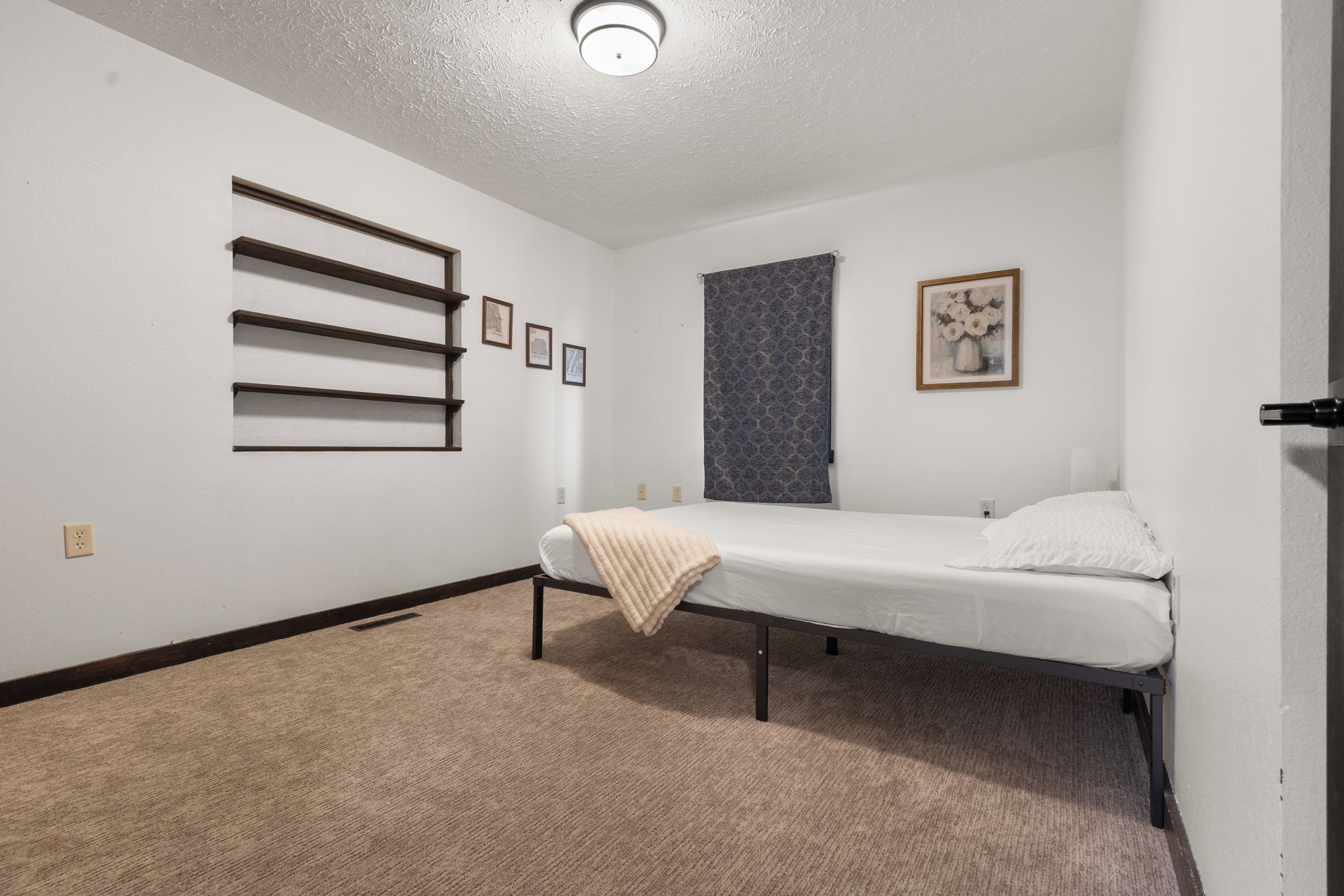 A single-bed room with a headboard, a bedside table, a wall-mounted shelf, a window with a patterned curtain, and a framed picture on the wall.