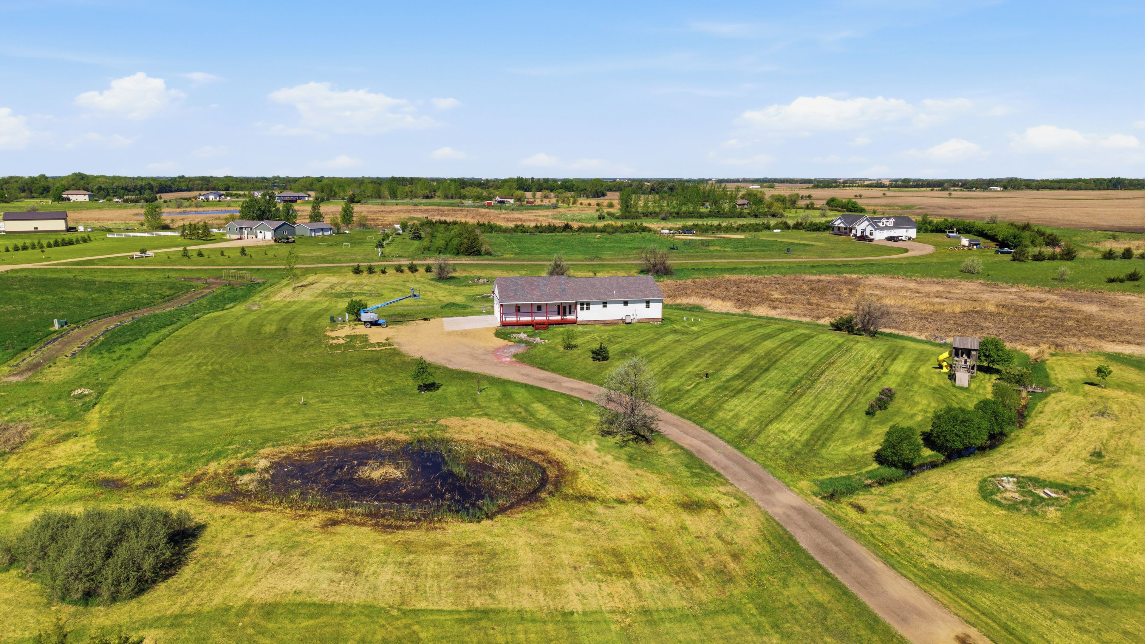 A rural landscape with a white house surrounded by green fields and a dirt path leading to it.