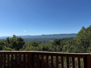 Uitzicht vanaf een houten terras op een bebost landschap met verre bergen, onder een heldere lucht.