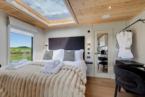A modern bedroom with a skylight, a large bed with fluffy bedding, a mirror, and a view of the outdoors.