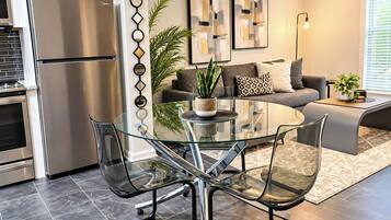 A modern kitchen with a stainless steel refrigerator, a glass dining table with chairs, and a living area with a sofa and decorative wall art.