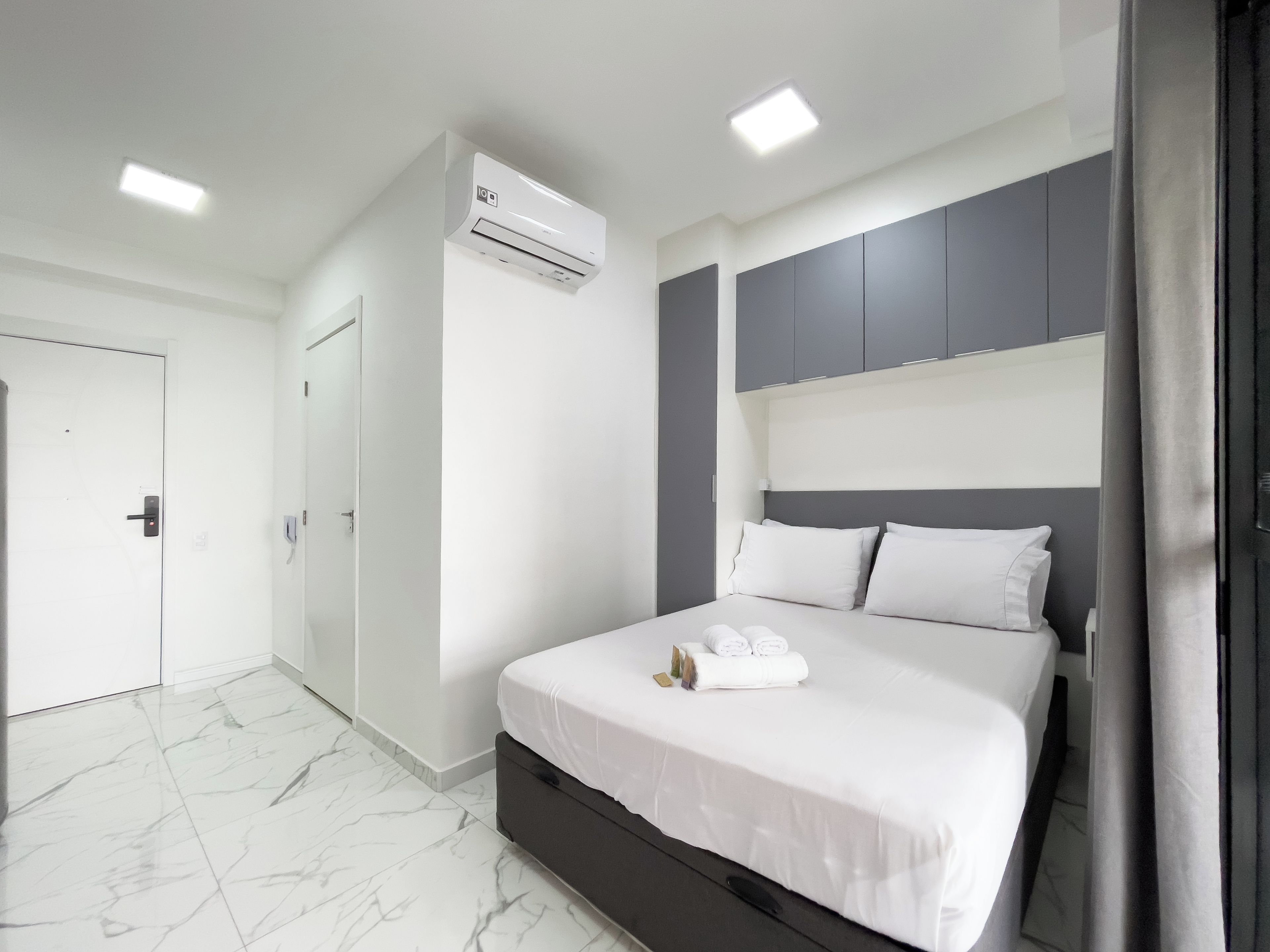 A modern hotel room with a bed, a wardrobe, an air conditioning unit, and a door.