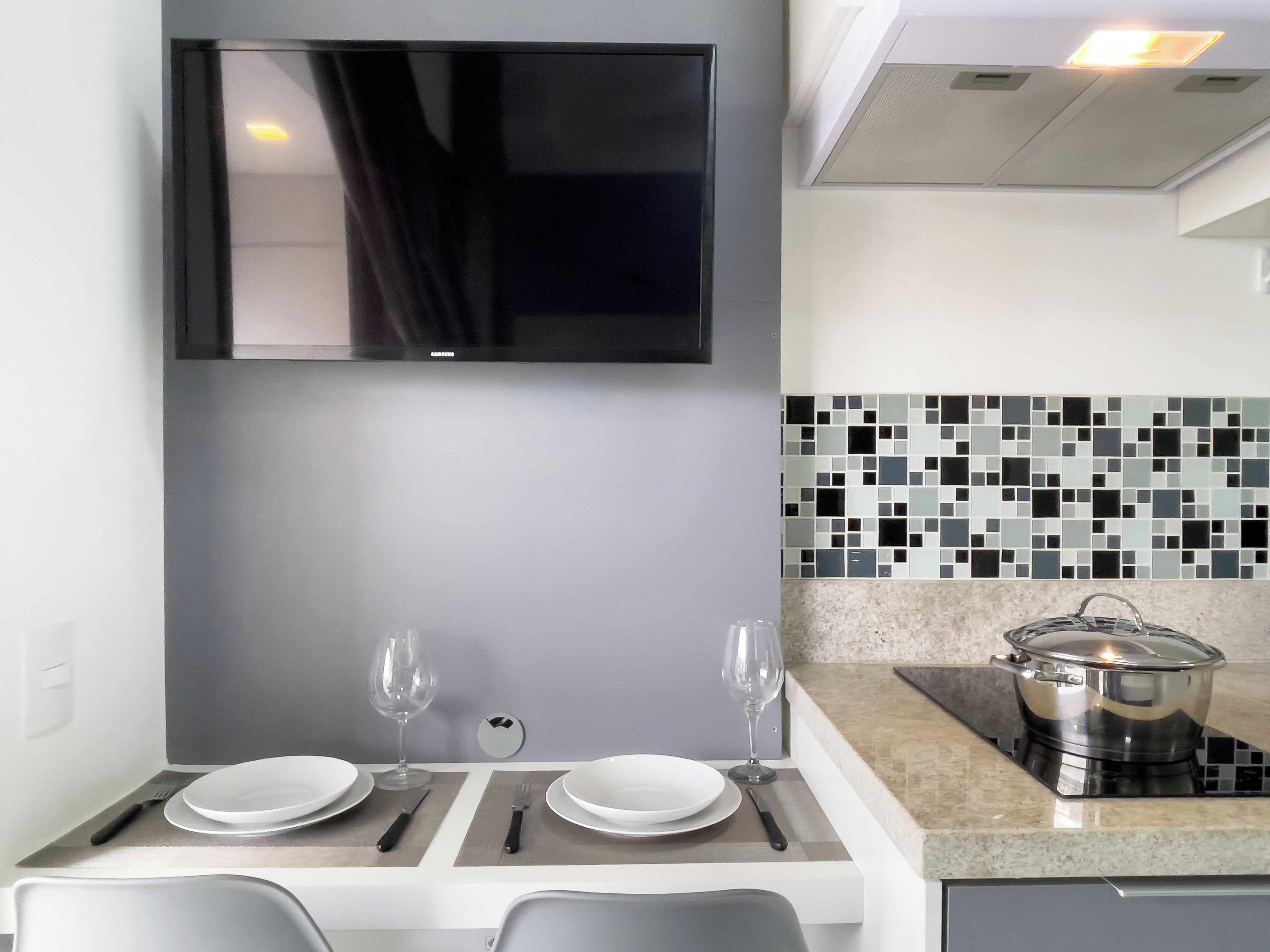 Una cocina moderna con un televisor de pantalla plana montado sobre una mesa de comedor para dos, con un salpicadero de azulejos y una olla de acero inoxidable sobre la encimera.