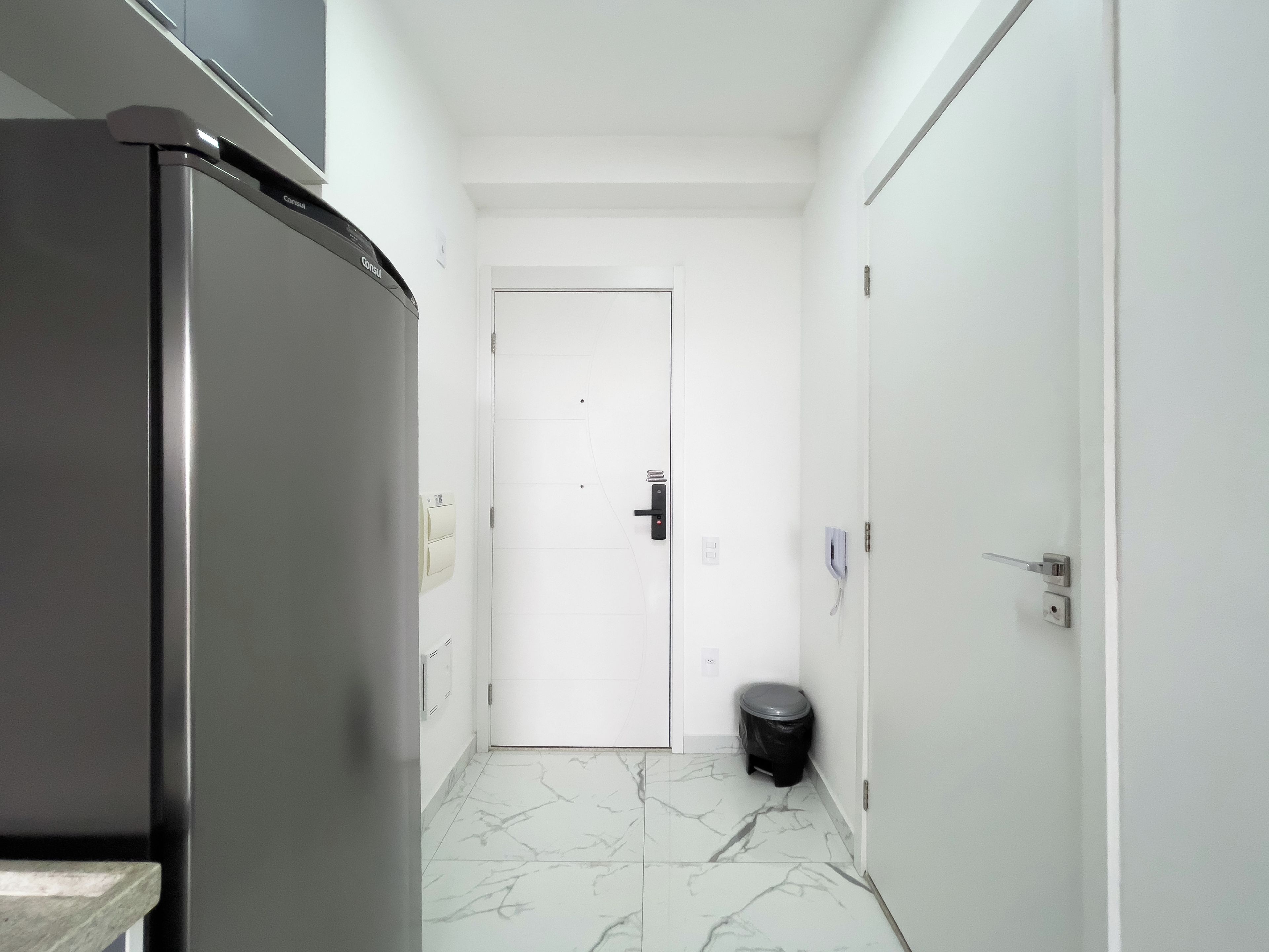 A narrow hallway with a refrigerator on the left, a door at the end, and a trash bin to the right.