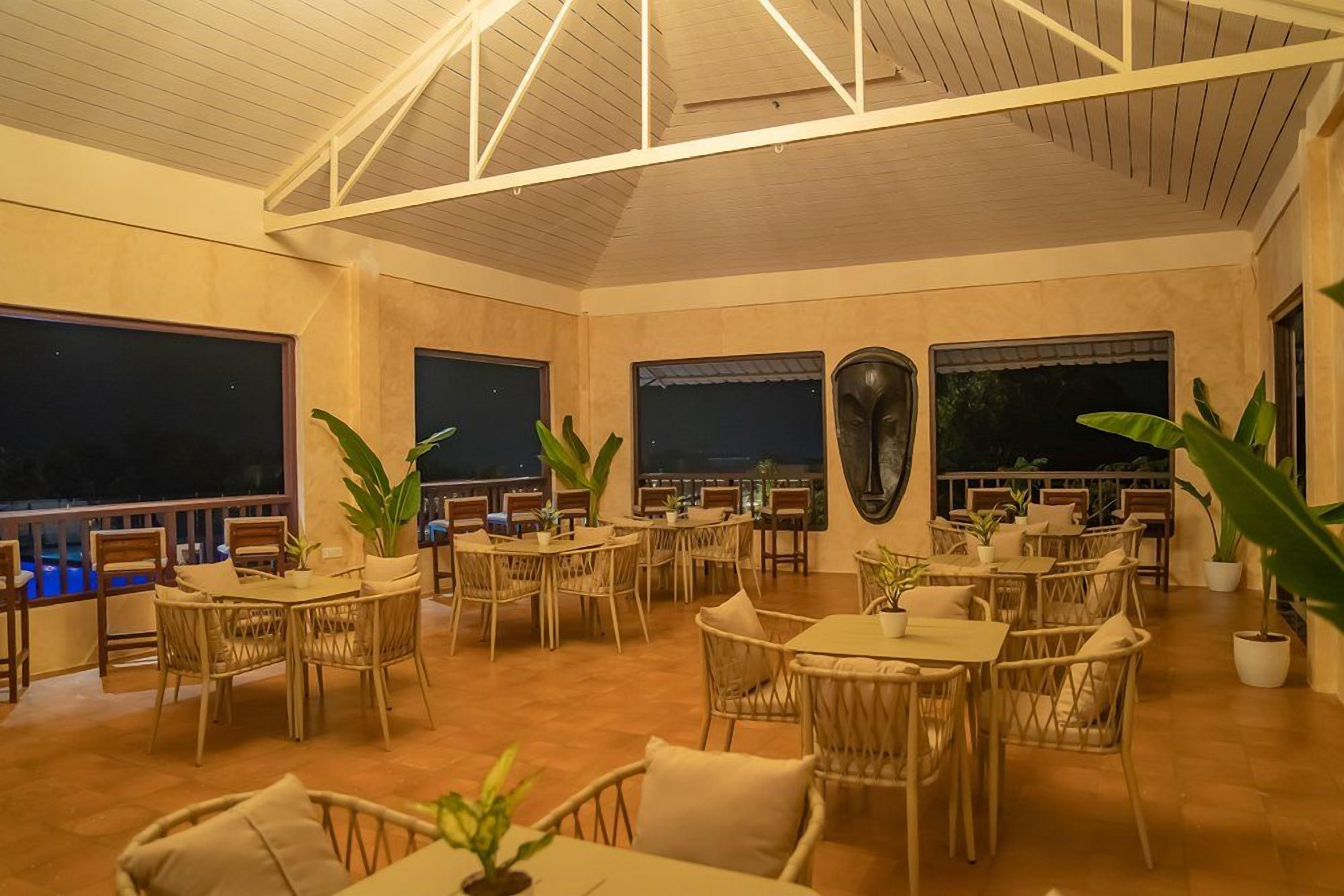 An indoor dining area with multiple tables and chairs, large windows, and potted plants.