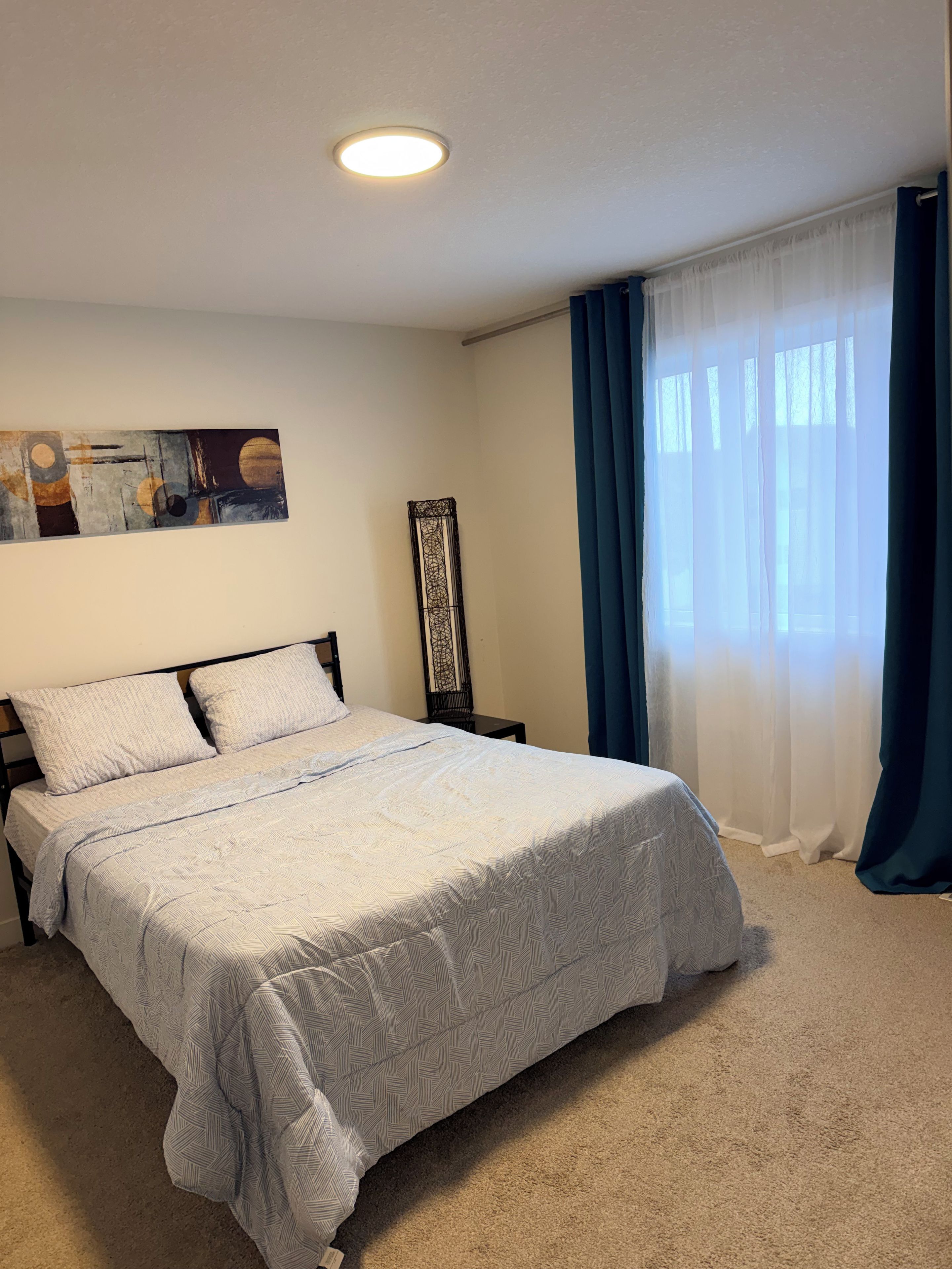 Ein Schlafzimmer mit einem Bett, einem Nachttisch, einem Wandbild und einem Fenster mit Vorhängen.