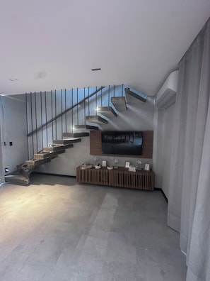 A modern living room with a staircase, a flat-screen TV mounted on the wall, and a wooden console table with decorative items. - New Listing (São Miguel dos Milagres)