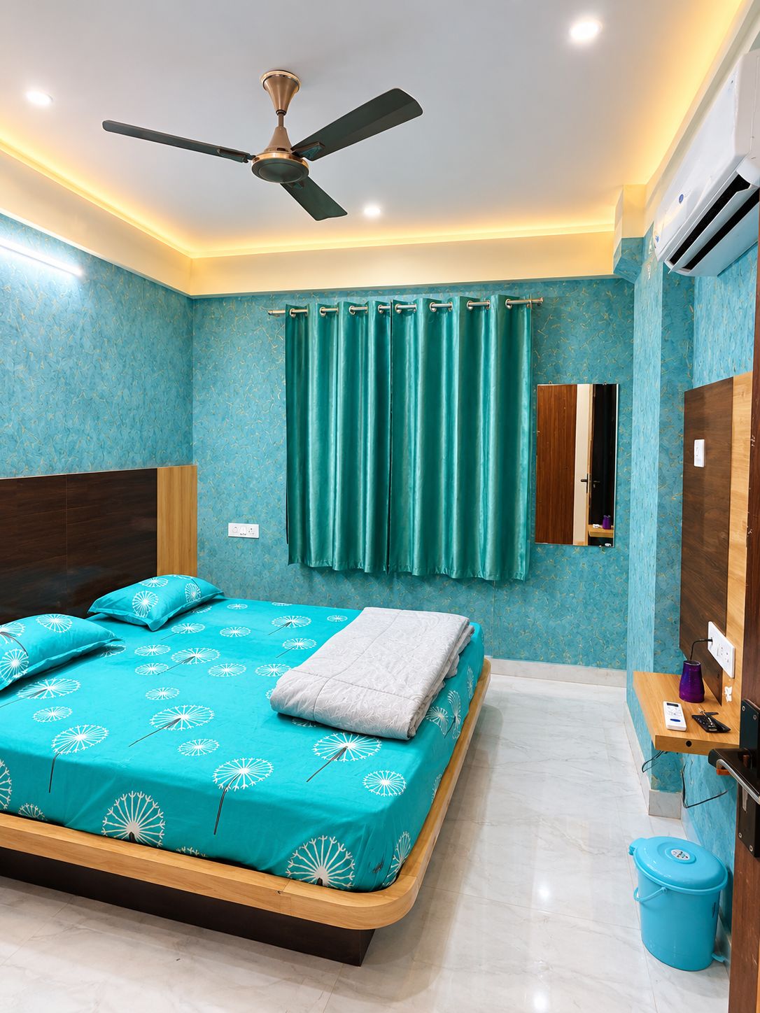 Un dormitorio con cama, ventilador de techo, cortinas color turquesa, espejo y un cubo de basura azul.