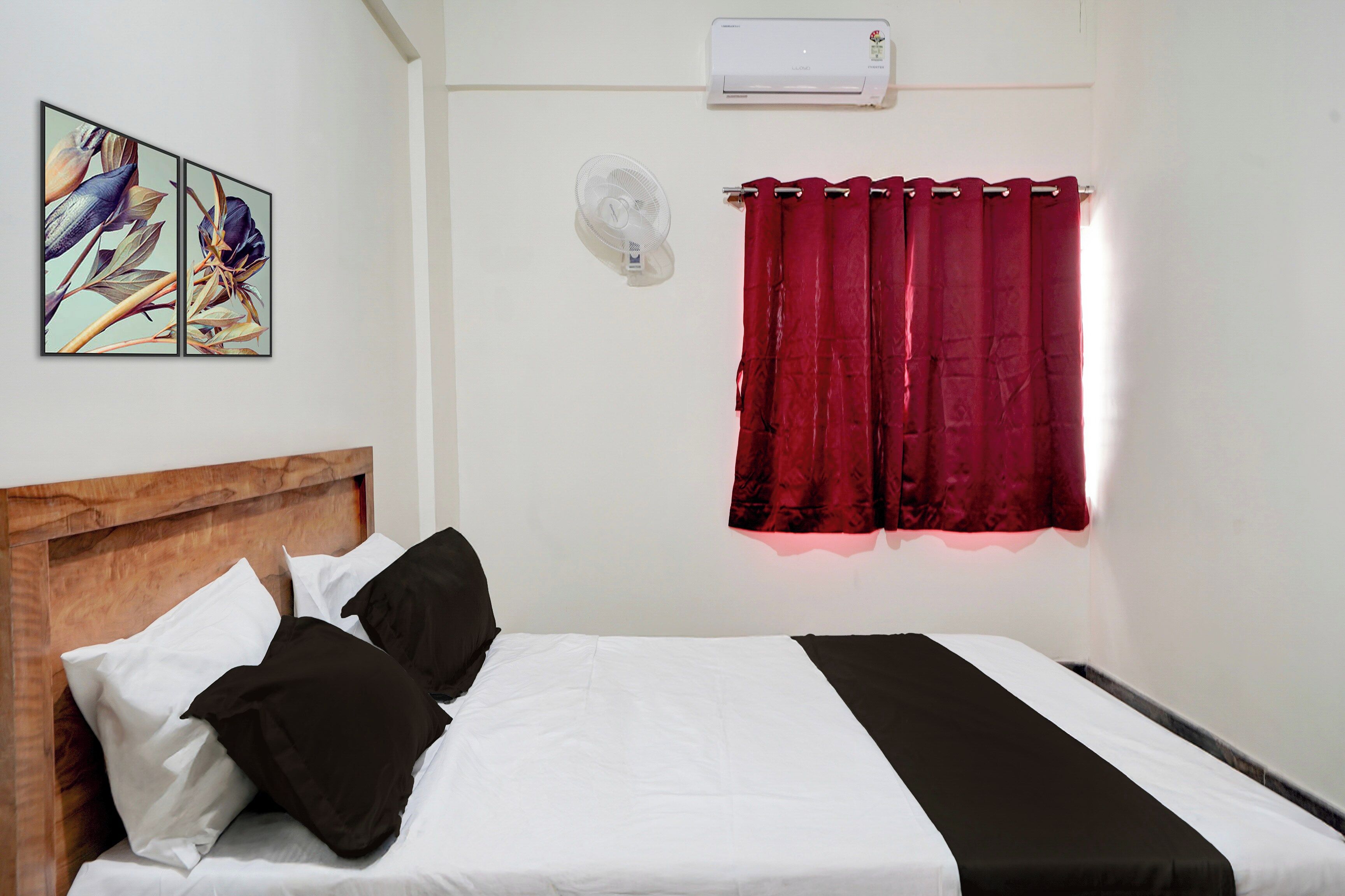 A bedroom with a bed, pillows, a headboard, a wall-mounted air conditioner, a window with red curtains, and a wall-mounted light fixture.