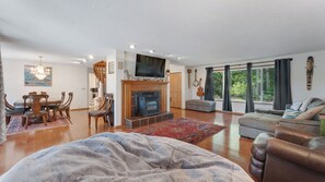 A spacious living room with a fireplace, a dining area with chairs, a sofa, and a large window with curtains. - Wooded Lakefront Retreat (Port Orford)