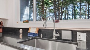 Una cocina con un lavamanos de acero inoxidable, una ventana con vistas a árboles, y un juego de cuchillos sobre la encimera.