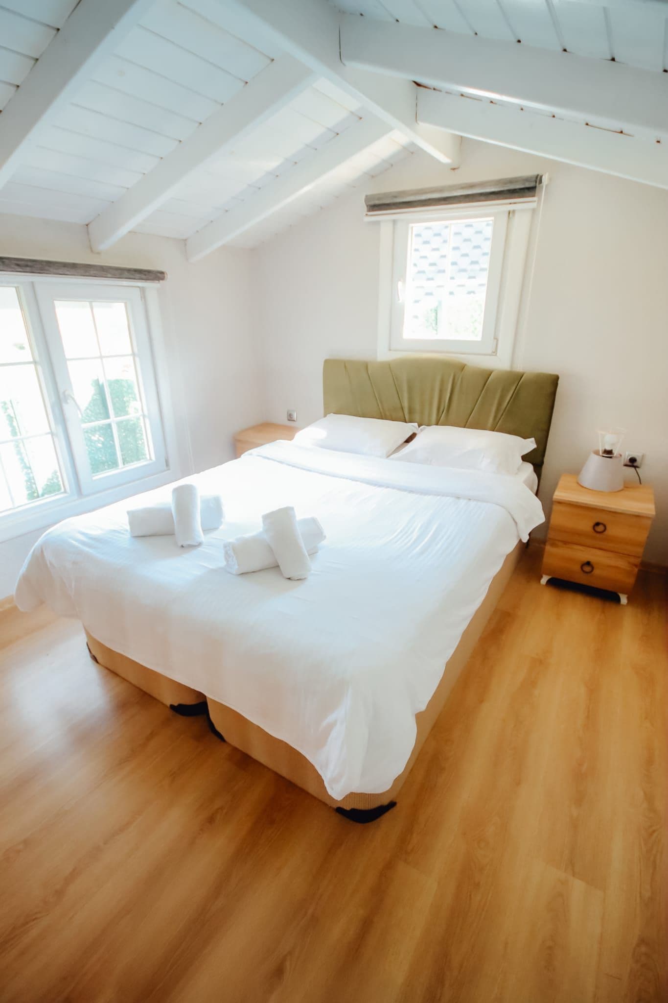 Una cama bien hecha con sábanas y toallas blancas, una mesita de noche de madera con una lámpara, y una ventana con persianas.