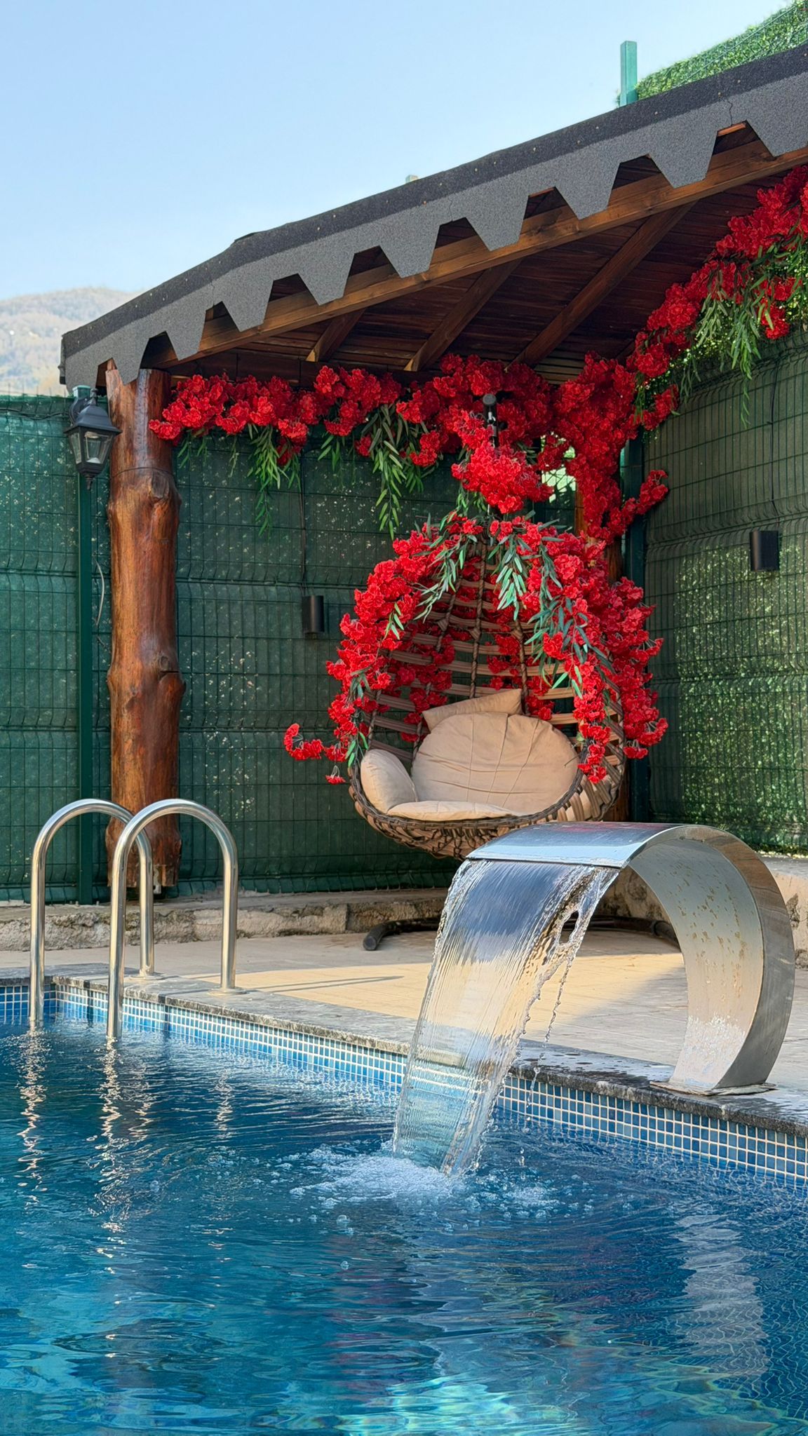 A swimming pool with a waterfall feature, a hanging chair, and red flowers.