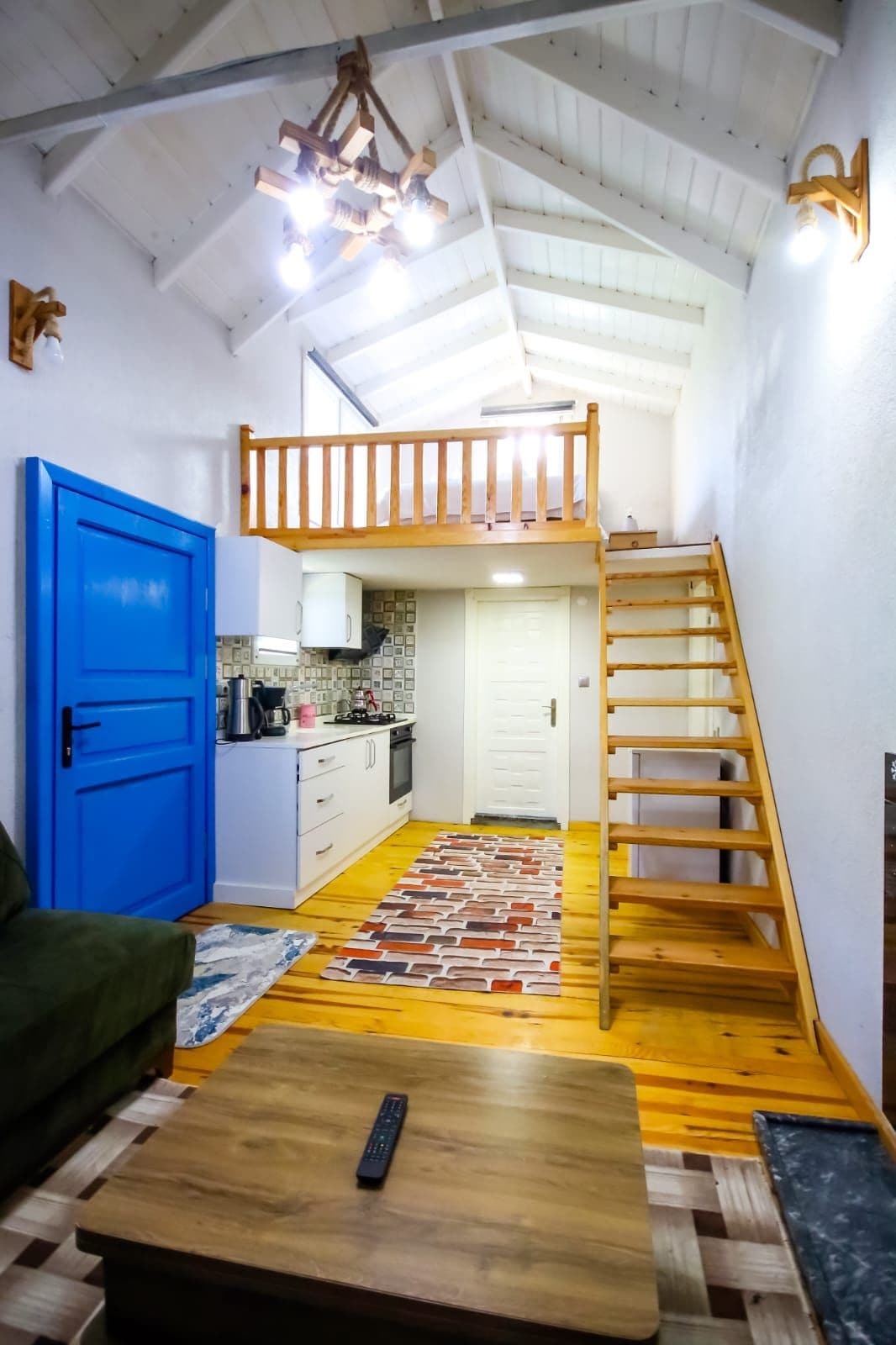 Un espacio habitable compacto con kitchenette, una puerta azul, una escalera de madera y un altillo.