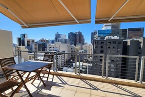 A rooftop terrace with a table and chairs, offering a view of a cityscape with modern buildings. - Manhattan Feeling! 2br, Huge Terrace & Epic Views, 2min Walk Bree Street (Cape Town)