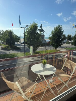 Un balcone con un tavolo in vetro e due sedie in vimini, affacciato su una strada con bandiere e una statua.