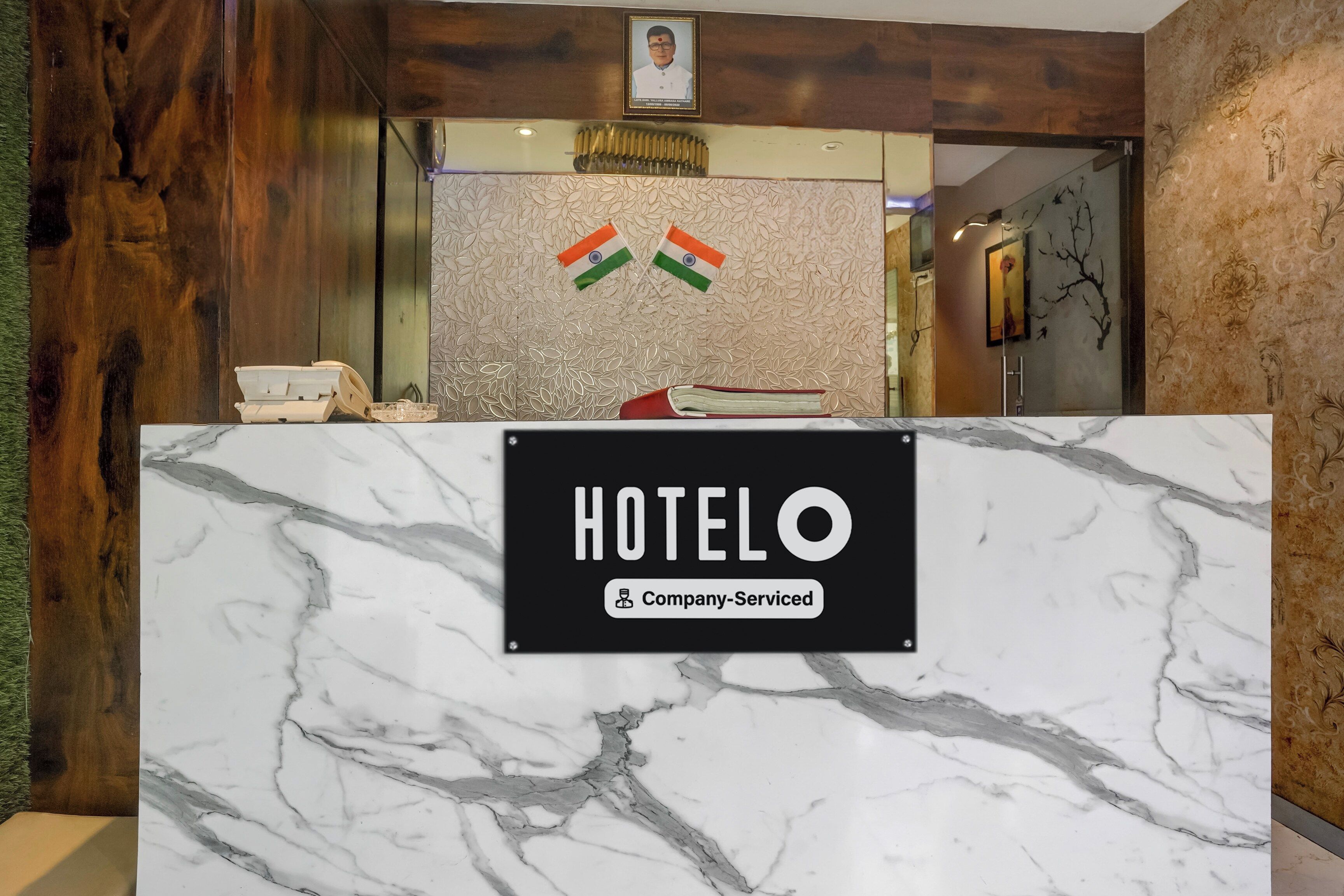 Un mostrador de recepción con encimera de mármol, un cartel que indica 'HOTEL' y 'Servicio de Empresa', y dos banderas de la India.