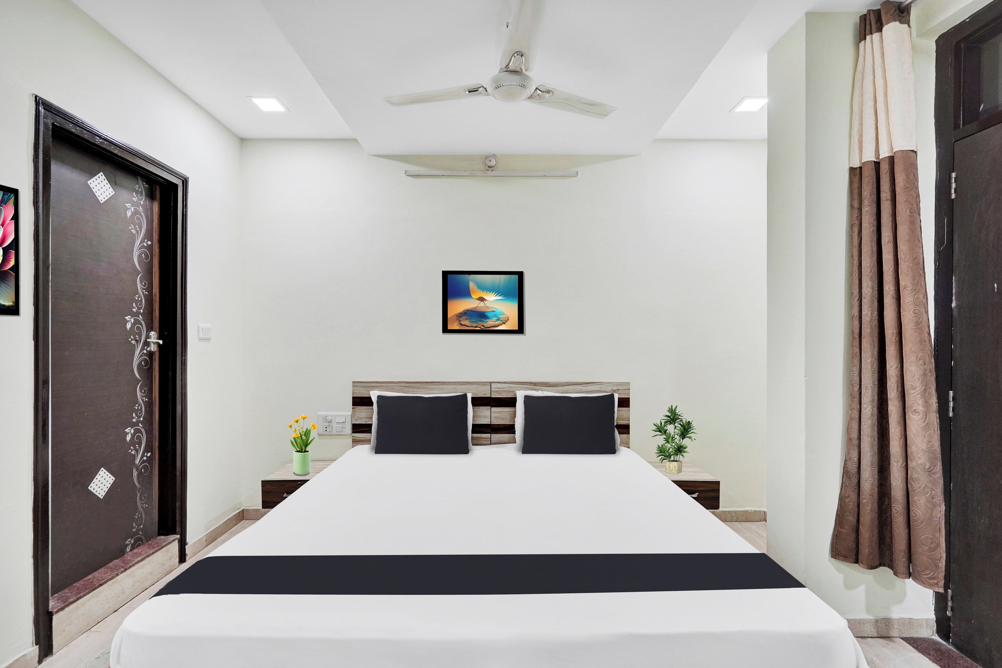 A hotel room with a bed, a ceiling fan, a painting, a nightstand, and a door.