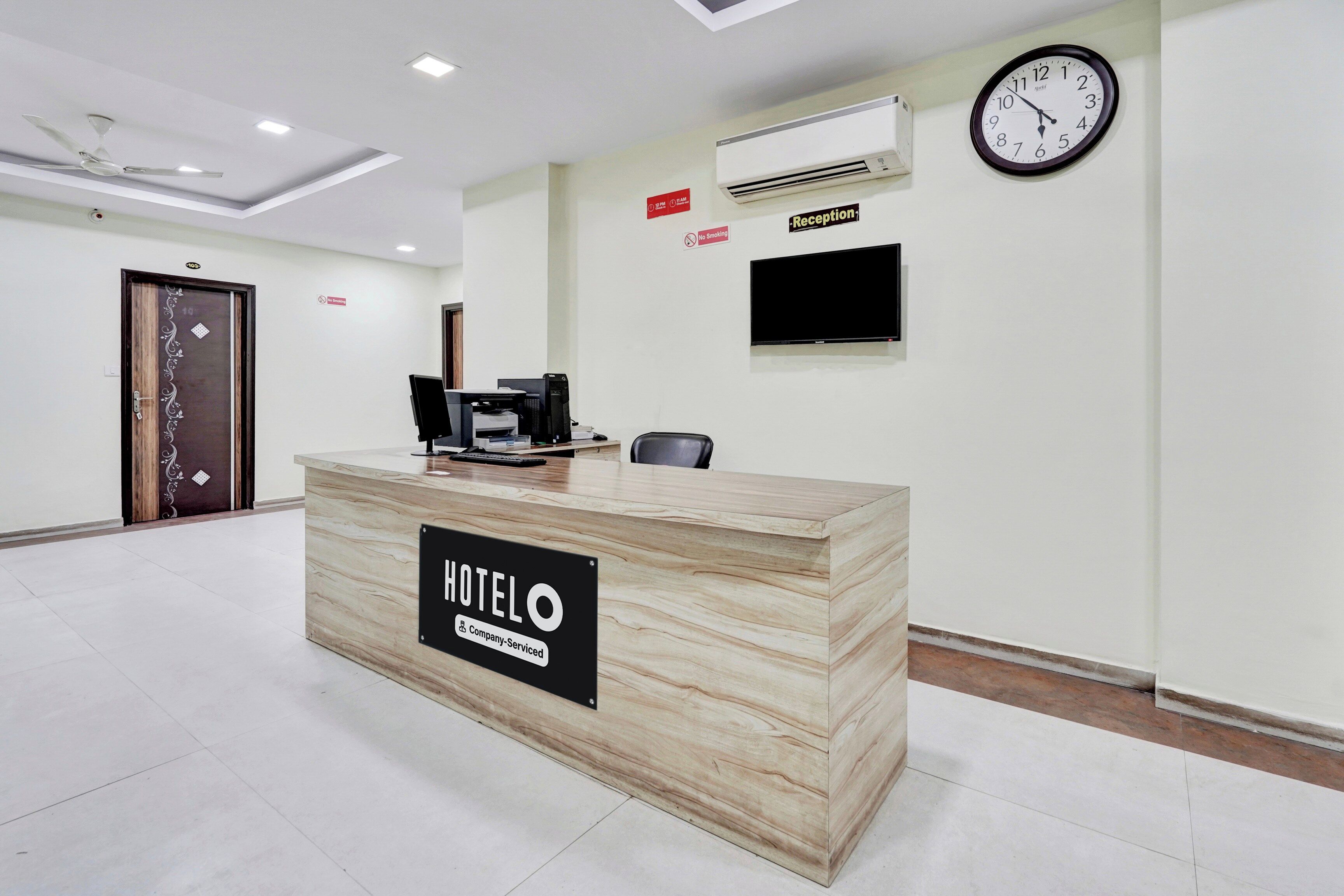 A hotel reception desk with a sign indicating it's company-serviced, a clock, and a no-smoking sign on the wall.