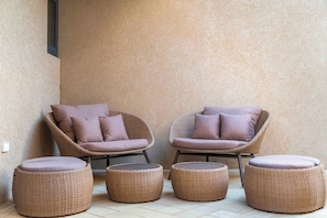 Two wicker lounge chairs with cushions, a small round table, and two ottomans on a tiled floor against a textured wall. - Ground-Floor 1BR Retreat | Oversized Private Patio (Noord)