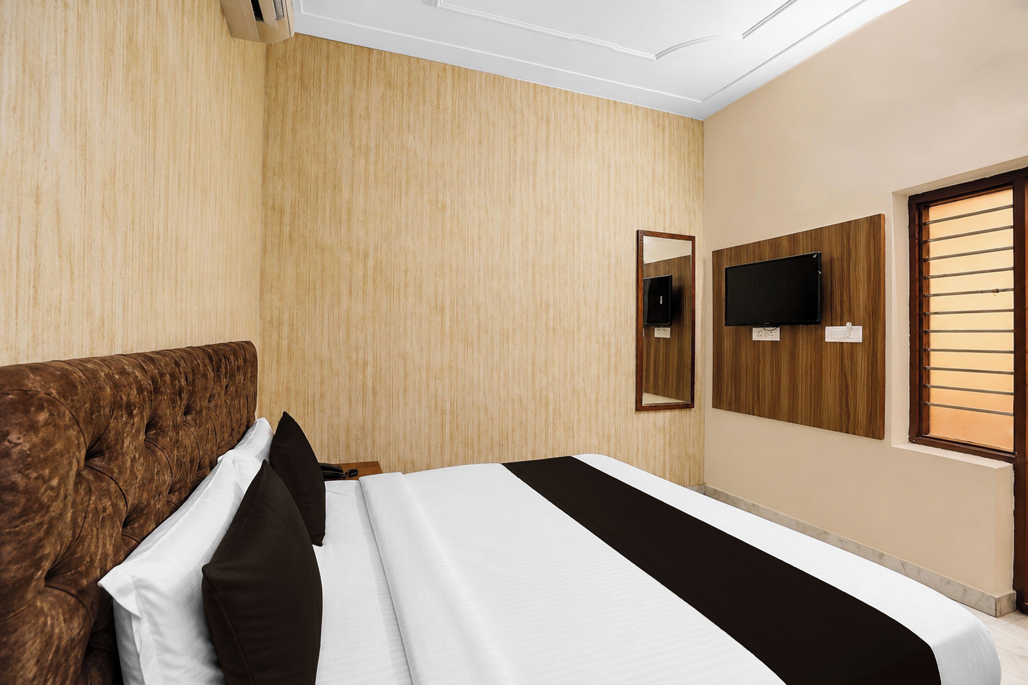 A hotel room with a large bed, a wooden headboard, a flat-screen TV mounted on the wall, and a window with horizontal blinds.