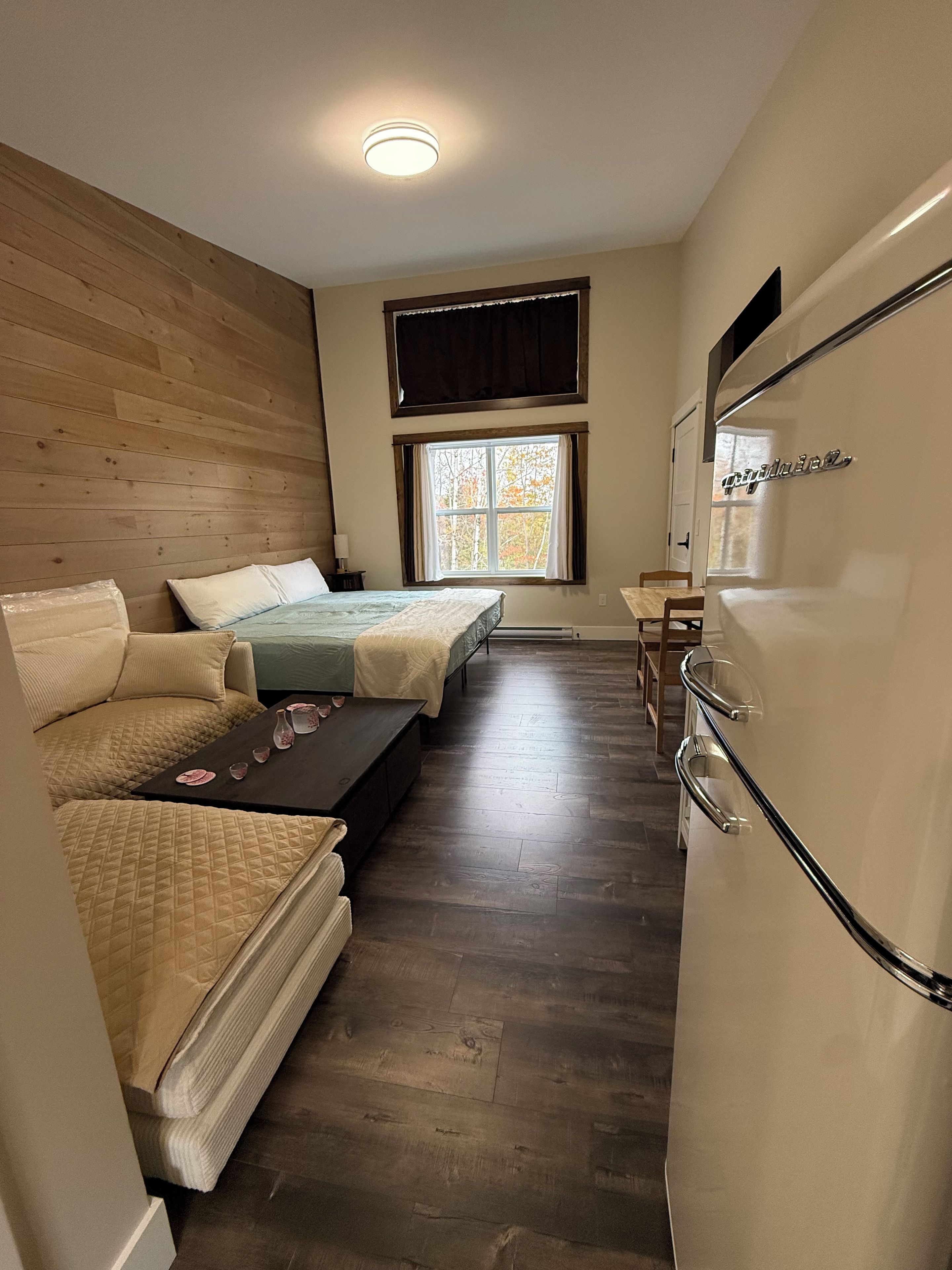 Ein kleines Zimmer mit zwei Betten, einem Tisch, einem Kühlschrank und einem Fenster mit Vorhängen.