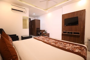 Habitación de hotel con cama, escritorio, silla, armario, televisor de pantalla plana montado en un panel de madera y unidad de aire acondicionado.