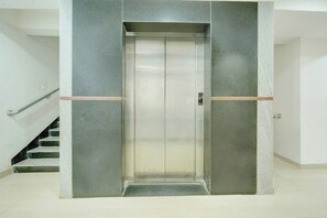 Un ascensor moderno con detalles metálicos, flanqueado por paredes grises y un pasamanos de metal a la izquierda.