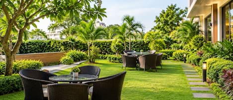 A well-maintained outdoor patio area with wicker furniture and a glass table, surrounded by lush greenery and a stone pathway.