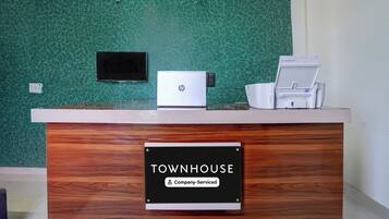 Mostrador de recepción con pared texturizada verde, un televisor de pantalla plana, una computadora portátil, una impresora y un letrero que indica 'TOWNHOUSE Company-Serviced'.