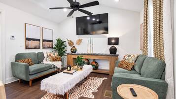 A modern living room with two green sofas, a white furry ottoman, a wooden coffee table, and a fireplace with decorative items.