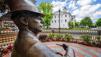 A bronze statue of a man in a hat pointing to the right, with a building and a fence in the background.