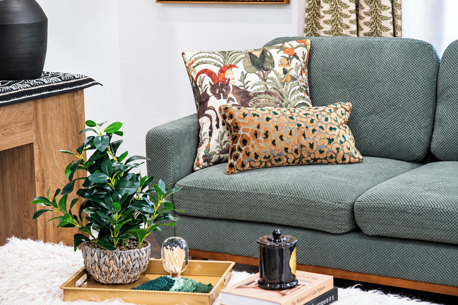 Ein Wohnzimmer mit einem grauen Sofa, einem gemusterten Zierkissen, einer Topfpflanze, einem dekorativen Glasbehälter und einem Buch.