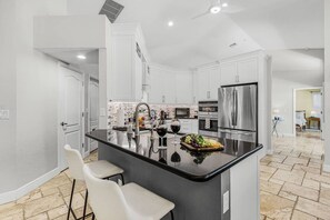 A modern kitchen with a central island, stainless steel appliances, and a seating area with white chairs. - Pineapple Paradise Retreat Private Pool and Spa (Bonita Springs)