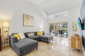 A modern living room with a grey sectional sofa, a wooden side table, and a large window providing natural light. - Pineapple Paradise Retreat Private Pool and Spa (Bonita Springs)