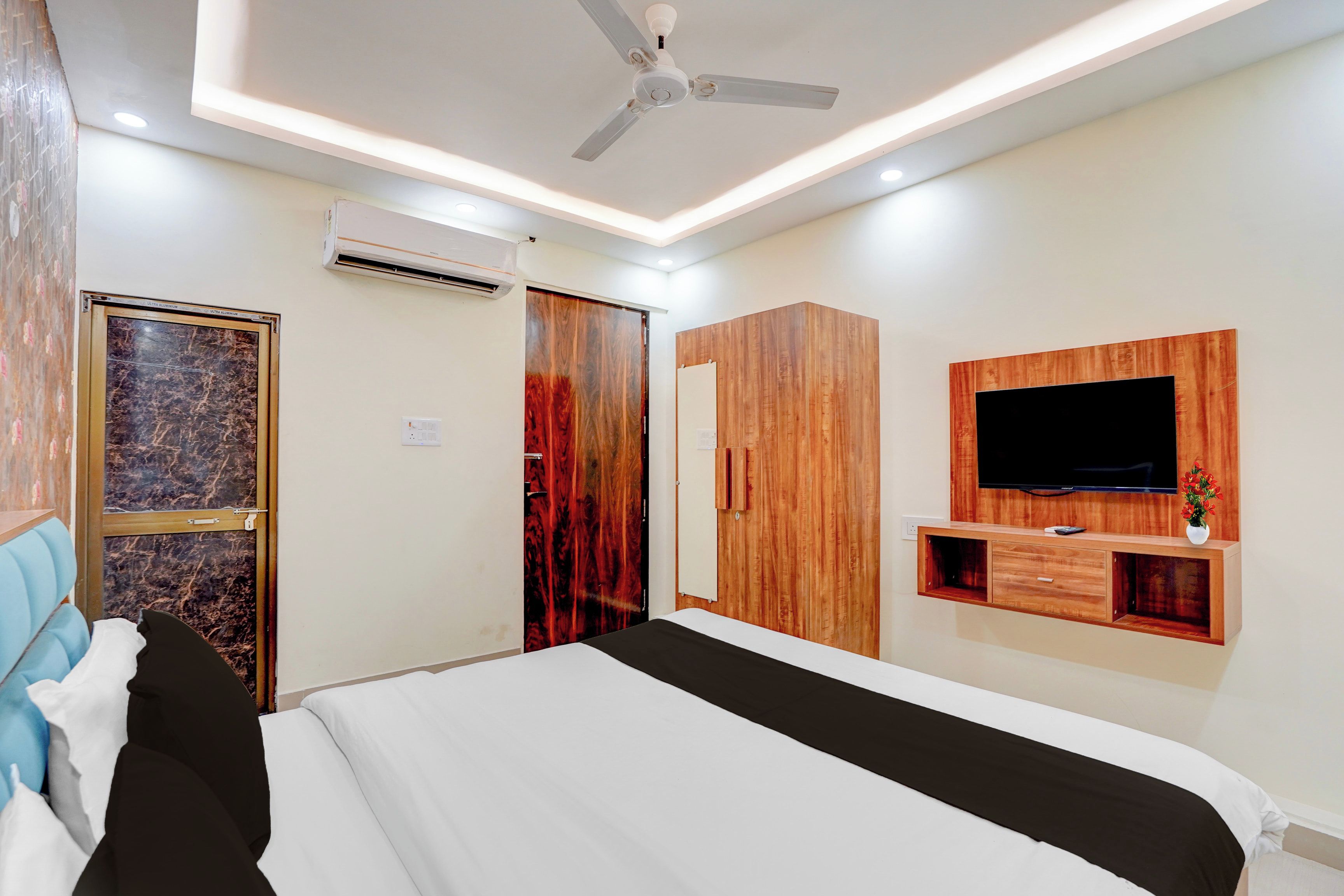 Una habitación de hotel con una cama, un ventilador de techo, un equipo de aire acondicionado, un armario de madera, un televisor montado en la pared y un pequeño estante con un florero.