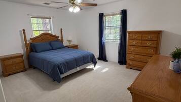 Une chambre à coucher comprenant un lit, une commode, une table de chevet, un ventilateur de plafond et une fenêtre avec des rideaux.